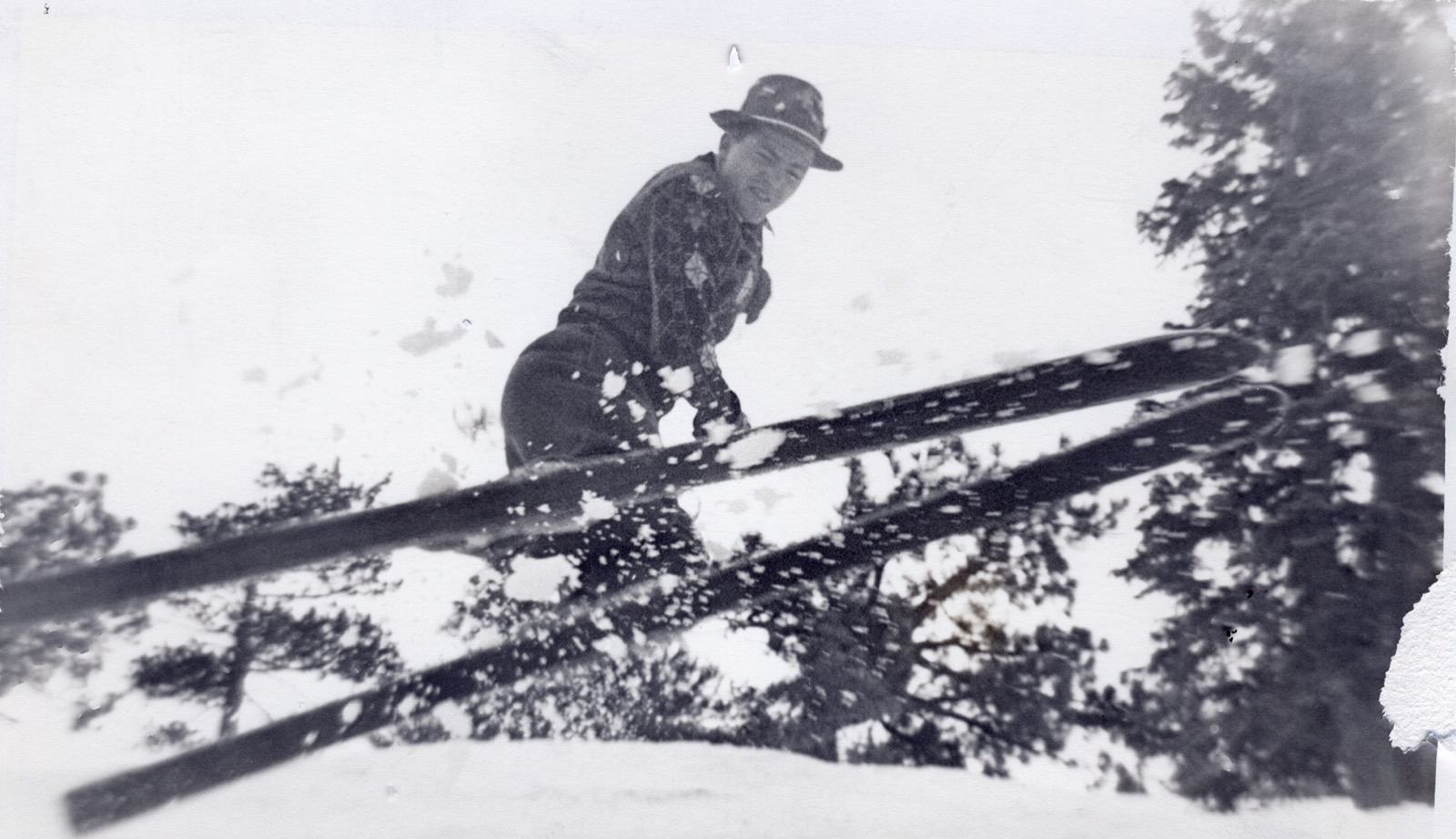 A young man in a patterned Fair Isle sweater and soft felt hat low on a snow slope, seen past a crossed pair of long wooden skis