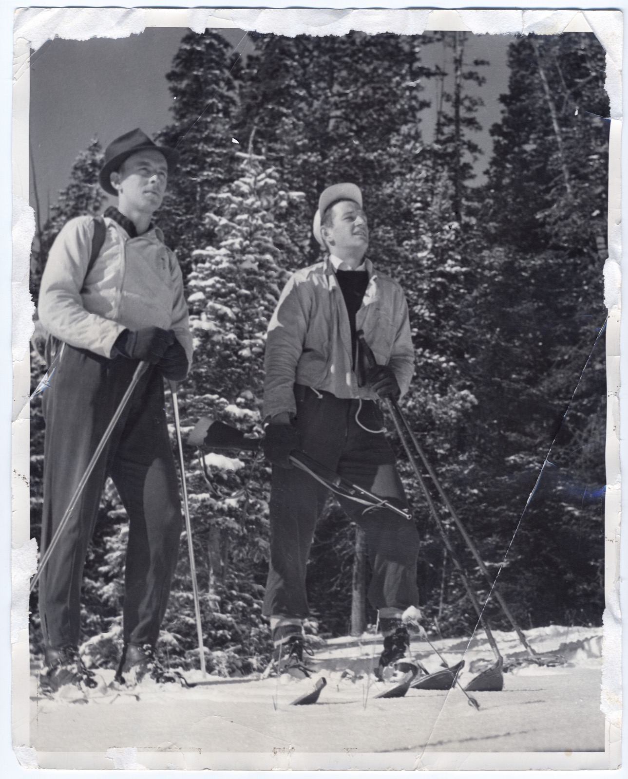 Two young men on skis in front of snow-laden conifers, one in a felt hat