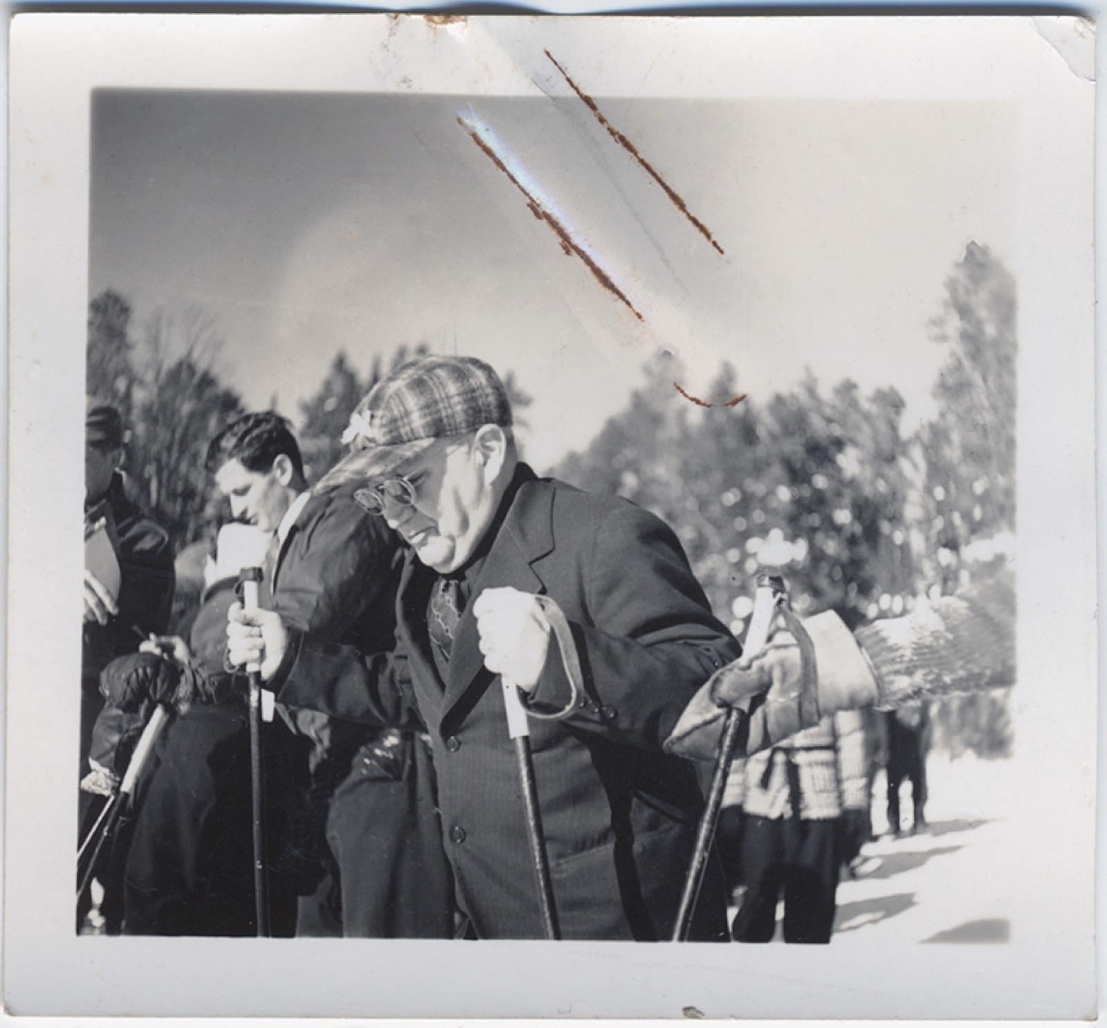 An older man in an overcoat, plaid cap and spectacles standing on skis in a crowd of skiers