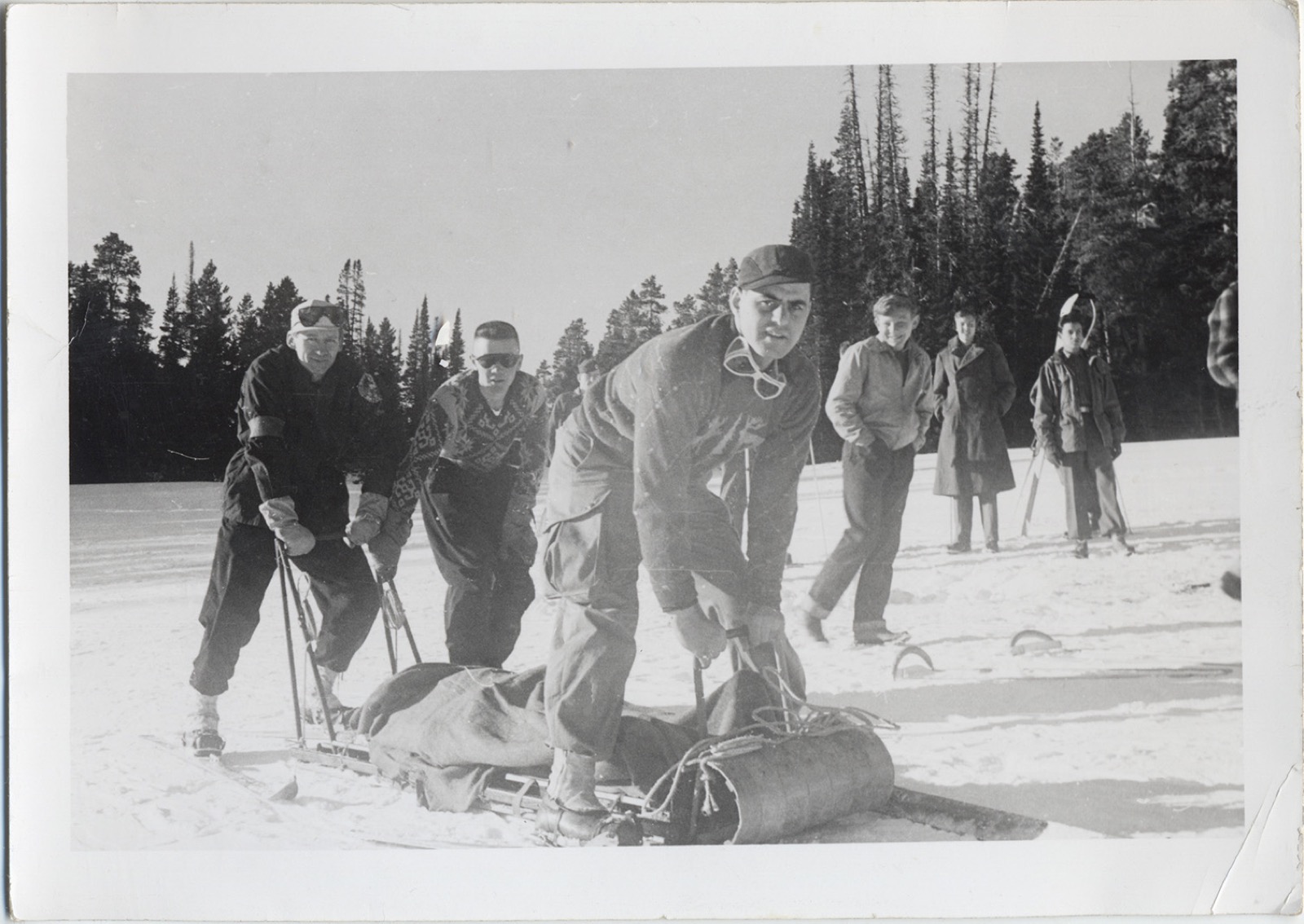 Three men handling a wooden rescue toboggan with rolled blankets and lashings, onlookers behind