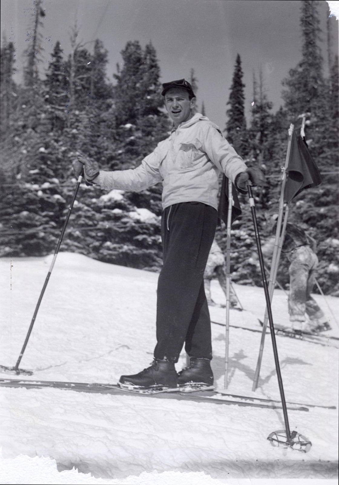 A young skier in a white anorak with a chest emblem, standing beside dark race flags with two skiers behind